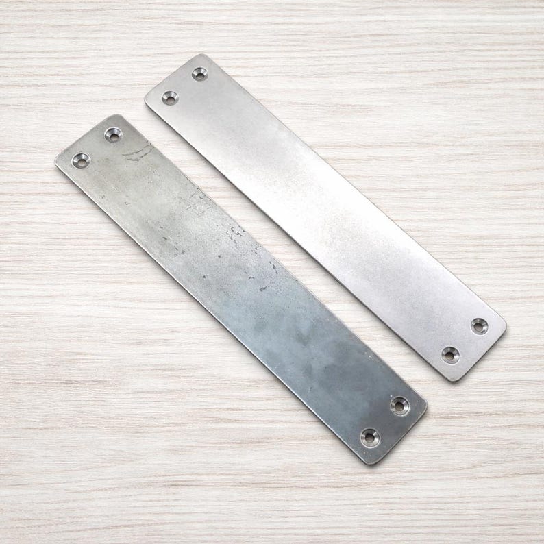 May include: Two rectangular metal plates with rounded corners and pre-drilled holes for screws. The plates are silver in color and have a brushed metal finish. They are lying on a light wood grain surface.