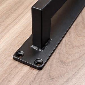 May include: Close-up of a black metal door pull handle. The handle is rectangular and welded to a flat plate with two mounting holes. The metal has a matte finish, and the background is a light wood grain.