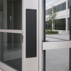 May include: A black rectangular door plate is mounted on a silver door frame. The plate is secured with four screws. The door is part of a commercial building with large glass windows.