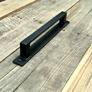 May include: A black metal door pull with a rectangular handle and a flat base. The handle is mounted on a weathered wooden surface. The door pull is designed for easy gripping and pulling. The hardware is likely for a cabinet or door.
