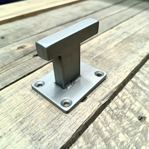 May include: A silver-colored metal bracket with a T-shaped design. The bracket has a rectangular base with four pre-drilled holes for mounting. The T-shaped top is solid and rectangular. The bracket is shown on a weathered wooden surface.