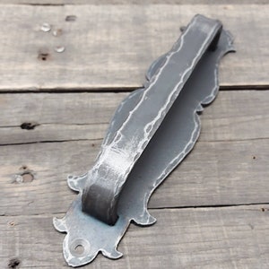 May include: A handcrafted, gray metal door pull with a hammered texture. The handle has a curved design and is attached to a decorative backplate with ornate edges. The hardware is designed for a rustic or vintage aesthetic.