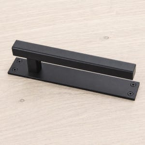 May include: A matte black door handle featuring a long, rectangular horizontal bar attached to a flat, rectangular base plate. The base plate includes four visible screw holes, one at each corner, with screws installed to secure the hardware.
