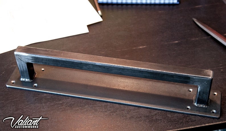 May include: A rectangular metal door pull with a welded handle. The handle is a square tube shape, and the base has pre-drilled holes for mounting. The metal has a dark, industrial finish. The logo "Valiant Customworks" is visible.