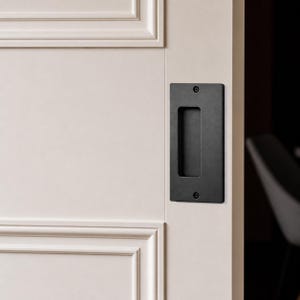 Peut inclure: Une poignée de porte encastrée rectangulaire noire installée sur une porte blanche. La poignée de porte a une découpe rectangulaire pour la prise et est fixée avec deux vis. La porte fait partie d'un mur à panneaux blancs.
