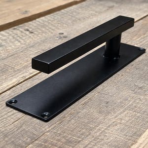 May include: A black door handle with a rectangular bar and a flat, rectangular backplate. The handle is mounted on a wooden surface. The handle is made of metal and has a modern, minimalist design.
