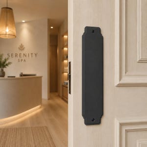 May include: A black door plate with decorative edges is mounted on a light-colored door. The door is partially open, revealing a spa reception area with the text "SERENITY SPA". A woven lampshade hangs from the ceiling.