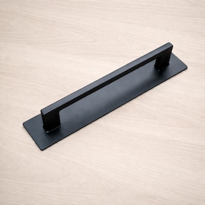 May include: A matte black door pull with a rectangular base and a horizontal bar handle. The handle is mounted on a rectangular plate, creating a modern, minimalist design. The hardware is likely made of metal.
