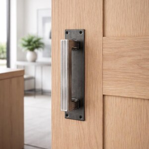 May include: A modern door handle with a rectangular, dark metal backplate and a brushed metal handle. The handle is mounted on a light-colored wooden door. The door is partially open, revealing a glimpse of a bright interior.