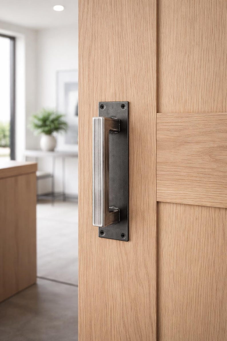 May include: A modern door handle with a rectangular, dark metal backplate and a brushed metal handle. The handle is mounted on a light-colored wooden door. The door is partially open, revealing a glimpse of a bright interior.