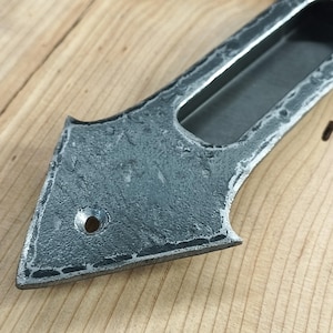 May include: Close-up of a hand-forged, gray metal drawer pull with a textured surface. The pull features a rectangular recessed handle and a decorative, pointed end. The metal has a rustic, aged appearance, with a small hole near the pointed end.