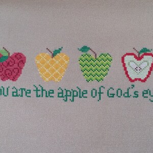 Puede incluir: Bordado de punto de cruz con cuatro manzanas rojas, amarillas y verdes y el texto "You are the apple of God's eye".