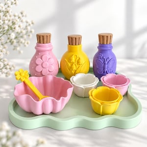 Puede incluir: Un juego de tres botellas coloridas con tapas marrones, un cuenco grande rosa con bordes festoneados y una cuchara amarilla con forma de flor, y tres cuencos pequeños con forma de flor en blanco, amarillo y rosa sobre una bandeja verde claro.