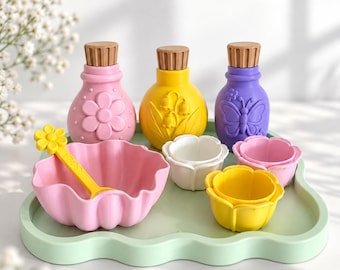 Puede incluir: Un juego de tres botellas coloridas con tapas marrones, un cuenco grande rosa con bordes festoneados y una cuchara amarilla con forma de flor, y tres cuencos pequeños con forma de flor en blanco, amarillo y rosa sobre una bandeja verde claro.