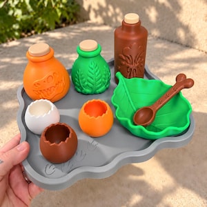 Puede incluir: Una bandeja de silicona gris que contiene varios juguetes: una botella naranja con relieve de dinosaurio, una botella verde con textura de hoja, una botella marrón, un cuenco verde con forma de hoja, una cuchara marrón y tres cáscaras de huevo rotas en blanco, naranja y marrón.