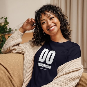 May include: A navy blue long-sleeve shirt with the words "DO SOMETHING" in white block letters. The text "Michelle Obama" is below the main text. The shirt is worn with a beige cardigan.