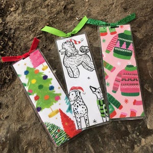 May include: Three Christmas-themed bookmarks with ribbon ties. One bookmark features a poodle wearing a Santa hat, another features a dalmatian wearing a Santa hat, and the third features a pink background with a green sweater with a Christmas tree design.