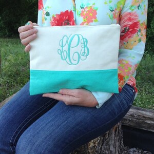May include: Cream-colored canvas clutch with a turquoise leather bottom and a turquoise monogram. The clutch has a zipper closure and is held by a person wearing a floral shirt and blue jeans.