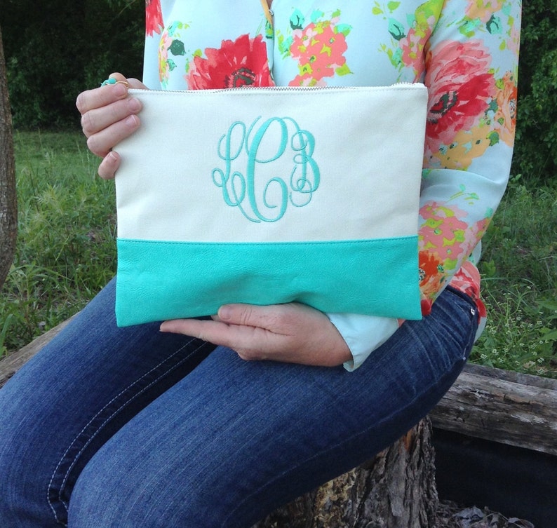 May include: Cream-colored canvas clutch with a turquoise leather bottom and a turquoise monogram. The clutch has a zipper closure and is held by a person wearing a floral shirt and blue jeans.