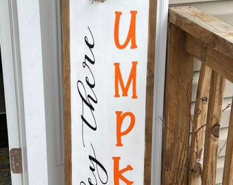 Könnte beinhalten: Ein weißes Holzschild mit einem braunen Holzrahmen. Das Schild sagt "Hey there Pumpkin" in schwarzer Kursivschrift. Das Wort "Pumpkin" ist vertikal mit orangefarbenen Buchstaben geschrieben. Das Schild ist mit einer Schleife aus Sackleinen und Grünzeug verziert.