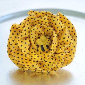 Puede incluir: Una flor de tela amarilla con lunares negros y un pequeño botón de abeja amarillo y negro en el centro.