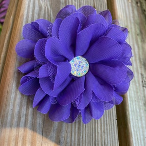 May include: A large purple flower with a sparkly rhinestone center. The flower is made of fabric and has a layered, ruffled design.