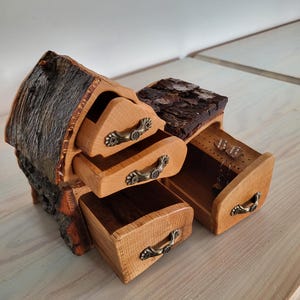 May include: A wooden jewelry box with a rustic bark exterior and three drawers. The drawers have ornate metal handles and are open to reveal the interior.