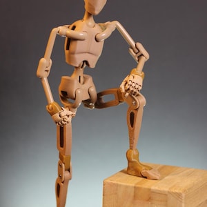 May include: A wooden articulated figure with a beige finish, standing on a wooden block. The figure is posed with one leg raised and the other leg straight. The figure has a detailed skeletal structure and is designed for use in art and animation.