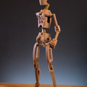 Puede incluir: Una figura articulada de madera con un acabado marrón. La figura está en posición de pie con los brazos doblados y las piernas ligeramente separadas. La figura tiene una cabeza, un torso, brazos y piernas, todos conectados por articulaciones. La figura está etiquetada como "DIY RIG" en el torso.