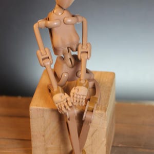 Peut inclure: Une figurine articulée en plastique beige avec des articulations brunes est assise sur un bloc de bois. La figurine est posée les jambes croisées et les bras croisés.