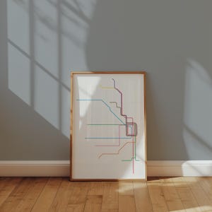 May include: A framed print featuring a colorful, minimalist subway map design. The map uses various colored lines, including blue, pink, green, and yellow, against a white background. The frame is a light brown color, and the print is leaning against a gray wall.