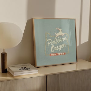 May include: A framed print with a vintage style illustration of the state of Oregon with a deer silhouette and the text "Portland Oregon Old Town". The print has a light blue background with a gold frame.