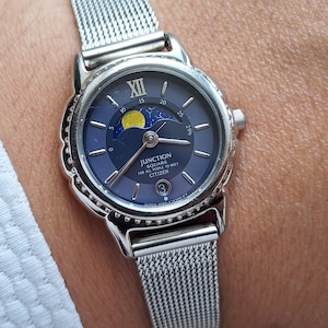 Könnte beinhalten: Eine silberfarbene Armbanduhr mit einem Mesh-Armband und einem dunkelblauen Zifferblatt. Das Zifferblatt zeigt eine Mondphasenanzeige und den Text "JUNCTION SOLAR CITIZEN". Die Uhr wird am Handgelenk getragen.