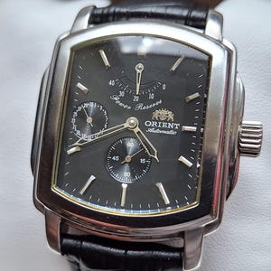 Può includere: Un orologio automatico Orient argentato di forma rettangolare con quadrante nero e cinturino in pelle nera. L'orologio presenta due sotto-quadranti e le parole "Power Reserve" e "Orient Automatic" sono visibili. Presentato in una scatola nera.