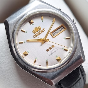 May include: Silver Orient Crystal 21 Jewels watch with a white face, gold detailing, and a black leather strap. The watch face shows the day and date. The watch is presented in a white box.
