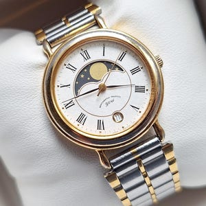 May include: A gold and silver wristwatch with a white face featuring Roman numerals and a moon phase indicator. The watch is in a white box with a black border, resting on a white pillow.