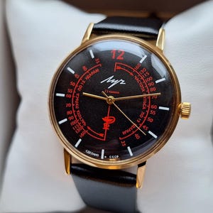 May include: A vintage gold-toned wristwatch with a black face and a black leather band. The watch face features red accents and the number 12 in red. The watch is presented in a black box with a white pillow.