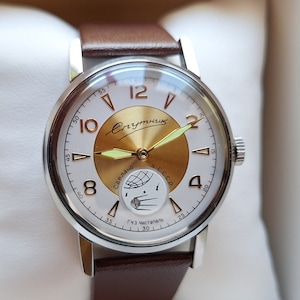 May include: A silver wristwatch with a brown leather band. The watch face is white with gold accents and features a moon phase indicator. The watch face also has the text "СССР" and "Ч.З. ЧАСОВЫЕ".