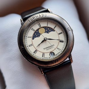 Könnte beinhalten: Eine Vintage-Quarzuhr "Tornado" mit braunem Lederarmband, präsentiert in einer weiß gepolsterten Box. Das Zifferblatt der Uhr zeigt römische Ziffern, ein Mondphasen-Design und eine Datumsanzeige. Das Uhrengehäuse hat eine Bronze-Oberfläche.