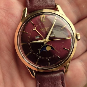 Può includere: Un orologio da polso color oro con quadrante bordeaux intenso e cinturino in pelle bordeaux abbinato. Il quadrante presenta lancette color oro, un indicatore delle fasi lunari, finestre per giorno e mese e la scritta Henry London in oro.