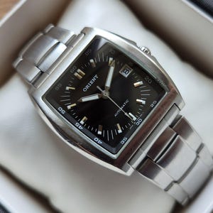Op de afbeelding: Een automatisch Orient-polshorloge met een rechthoekige roestvrijstalen kast en bijpassende schakelarmband. Het horloge heeft een zwarte wijzerplaat met zilverkleurige uurmarkeringen, lichtgevende wijzers en een datumvenster op de drie-uur-positie.