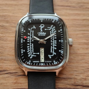May include: A black wristwatch with a square face and a black leather strap. The watch face has white markings and a red dot at the top. The watch face also has the text "СССР" and "СБТ 7" on it.