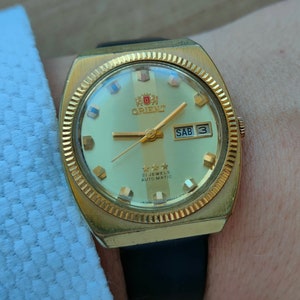May include: A gold-toned wristwatch with a white face and black leather band. The watch face has a date window that reads "SAB 3" and the text "21 JEWELS AUTOMATIC" is printed below the center of the face. The watch brand "ORIENT" is printed in black letters on the face.