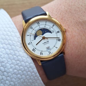 Può includere: Orologio da polso color oro con quadrante bianco, numeri romani e indicatore delle fasi lunari. L'orologio ha un cinturino in pelle blu navy. Il quadrante mostra il marchio "HENO QUARTZ" e la data.