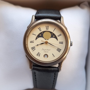 May include: A Jupiter 5 BAR wristwatch with a cream-colored face, Roman numerals, and a moon phase design. The watch has a black leather band and is presented in a white-padded box.