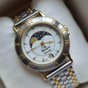 Puede incluir: Un reloj de pulsera Champion Quartz bicolor con esfera blanca, detalles dorados y correa plateada. El reloj presenta números romanos, un indicador de fase lunar y una pantalla de fecha. El reloj se presenta en una caja blanca.