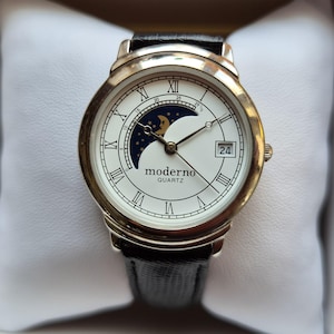 Könnte beinhalten: Eine silberfarbene Uhr mit weißem Zifferblatt, römischen Ziffern und Mondphasenanzeige. Die Uhr hat ein schwarzes Lederarmband und das Wort "moderno" ist auf dem Zifferblatt aufgedruckt. Das Datum wird angezeigt.