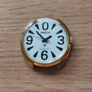 May include: A gold-tone wristwatch with a white face and black markings. The watch face has black numbers 3, 6, 9, and 12, and the words "PAKEТА" and "СДЕЛАНО В СССР".