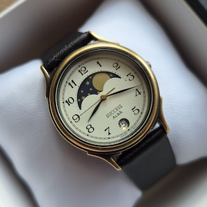 May include: A gold-toned Alba Success watch with a black leather band, displayed in a white box. The watch face is cream-colored with black numerals, gold hands, and a moon phase indicator. The watch is a classic timepiece.