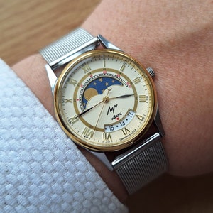 May include: A silver and gold wristwatch with a white face and a moon phase complication. The watch has a silver mesh band and a date window at the 4 o'clock position. The watch face also features the text "Сделано в СССР" which translates to "Made in USSR".